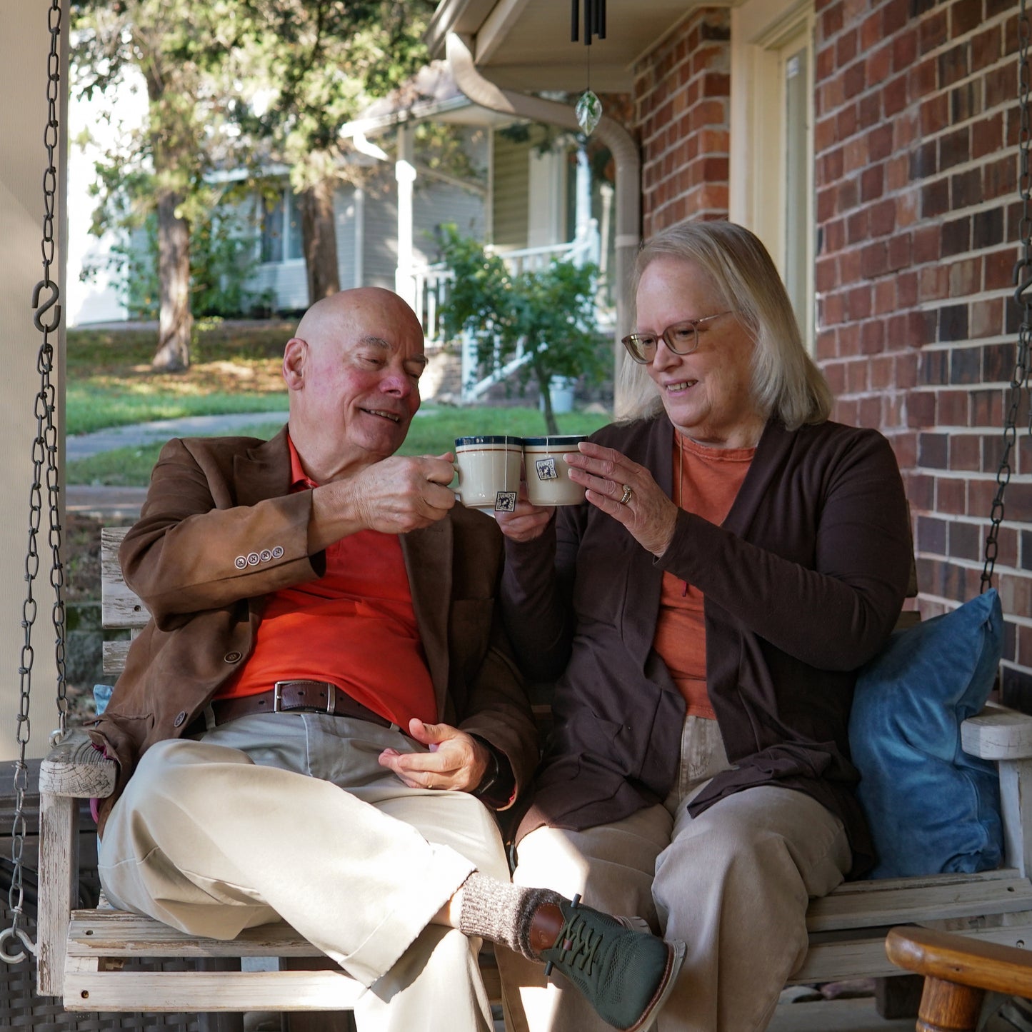 An elderly man and woman sit together on a porch swing, smiling and clinking mugs of Piper and Leaf Tea Co.'s Inflammation Relief blend, with trees and houses in the background.
