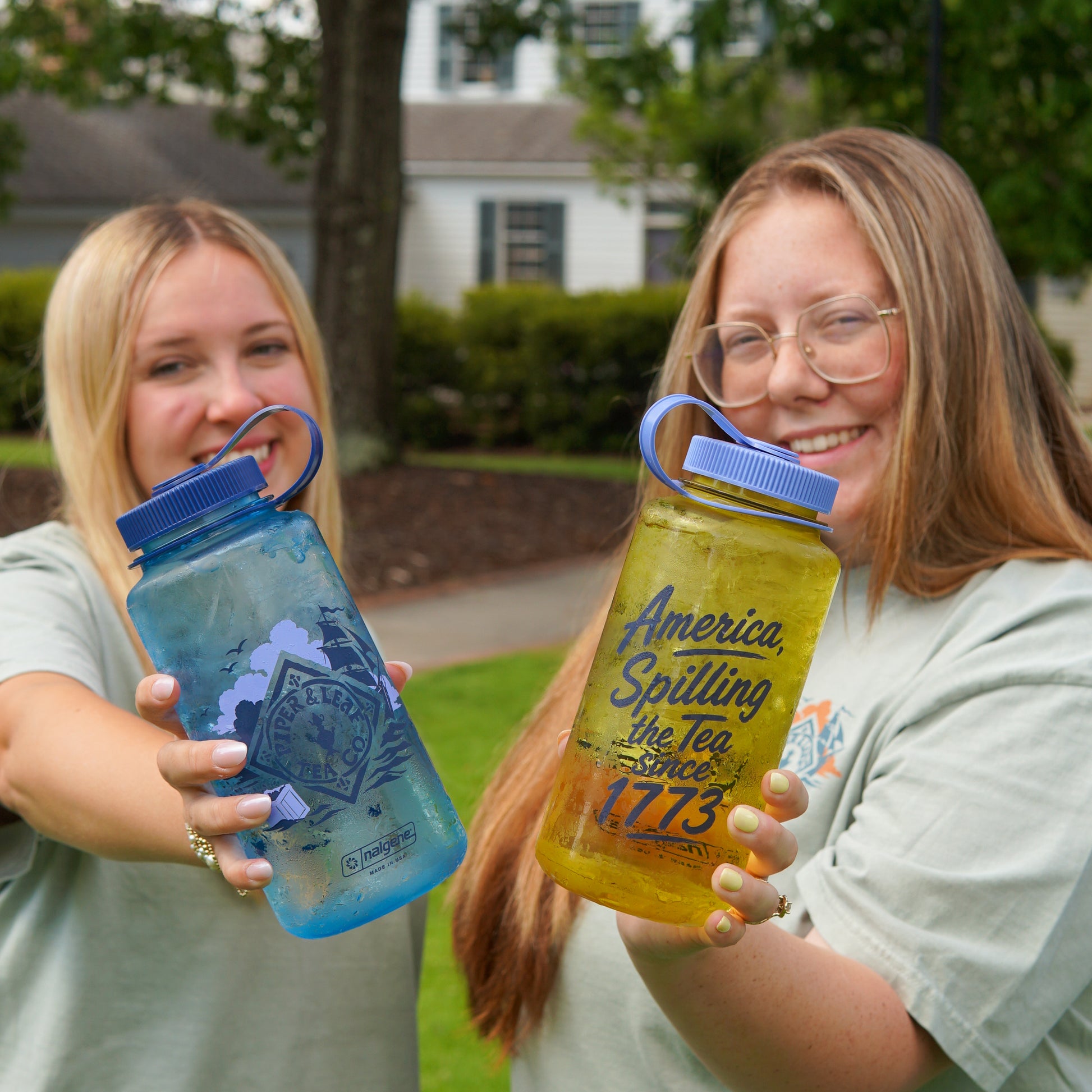 Two young women outdoors hold up Piper and Leaf Tea Co.'s "Spilling the Tea Nalgene" reusable water bottles, featuring tea-inspired designs and the phrase "America, Spilling the Tea Since 1773.