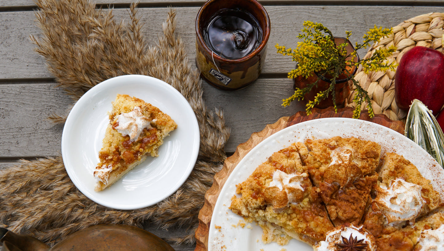 A crumb-topped pie slice with cream sits beside a Caramel Apple Pie Pound Bag (190 servings) by Piper & Leaf Tea Co., an iced glass of loose leaf tea, and autumn decor on a wooden table.