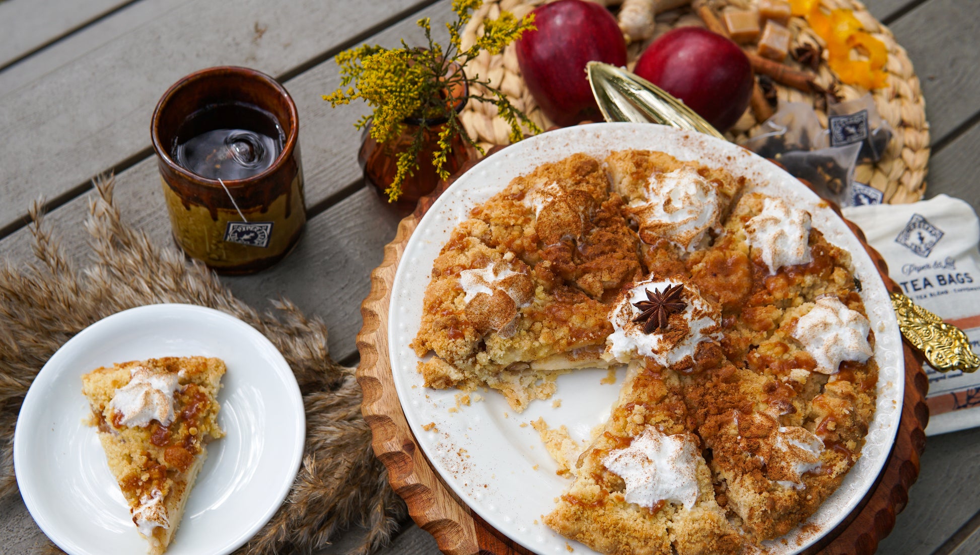 A round crumb-topped pie with cream sits on a platter, one slice served nearby. Piper & Leaf Tea Co.’s Caramel Apple Pie 9ct Tea Bags in Muslin, apples, honeycomb, and tea bags are arranged artfully on the wooden table.