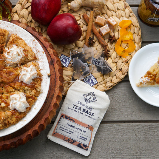 A slice of apple pie, whole apple pie, apples, caramel, cinnamon sticks, orange peel, and Piper & Leaf Tea Co. Caramel Apple Pie 9ct Tea Bags in Muslin are artfully arranged on a wooden table.