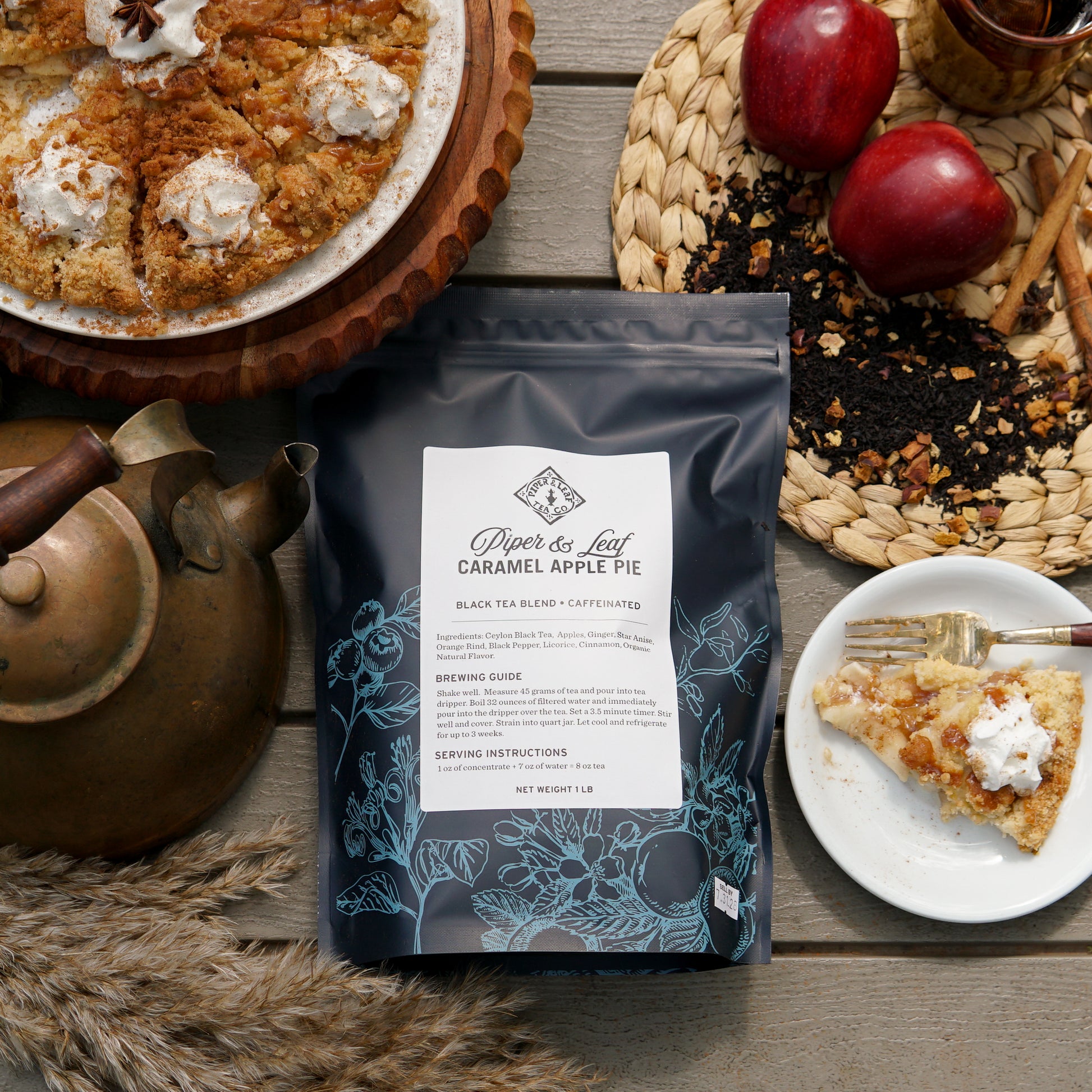 A Piper & Leaf Tea Co. Caramel Apple Pie Pound Bag (190 servings) sits on a table with a teapot, apple pie slice, two apples, Ceylon Black Tea loose leaf, and a woven placemat.