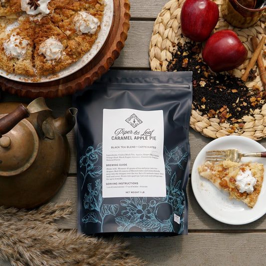 A Piper & Leaf Tea Co. Caramel Apple Pie Pound Bag (190 servings) sits on a table with a teapot, apple pie slice, two apples, Ceylon Black Tea loose leaf, and a woven placemat.