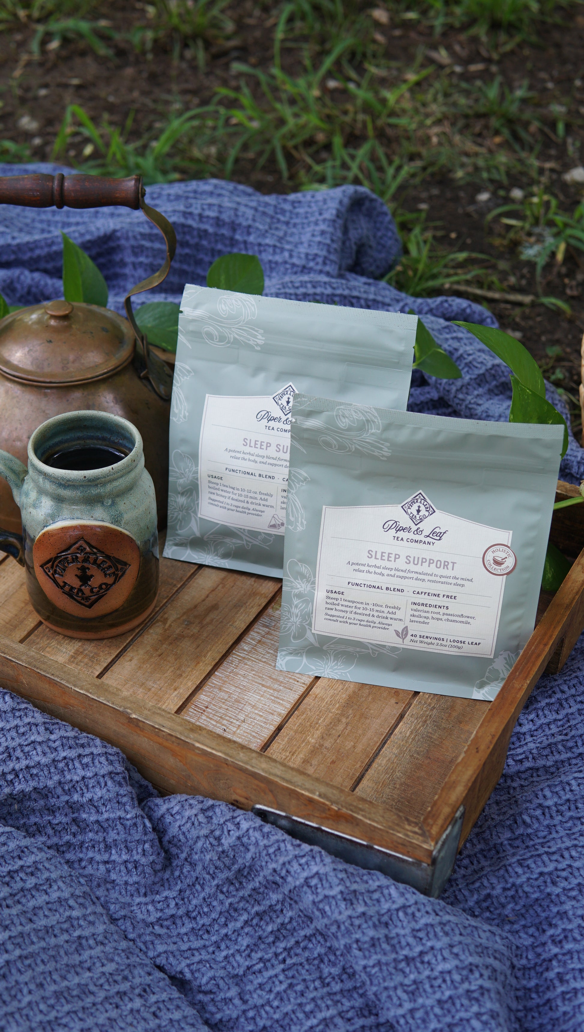 Two packages of Piper and Leaf Tea Co. Sleep Support - Holistic Collection tea, a ceramic mug, and a metal teapot sit on a wooden tray atop a blue textured blanket outdoors.