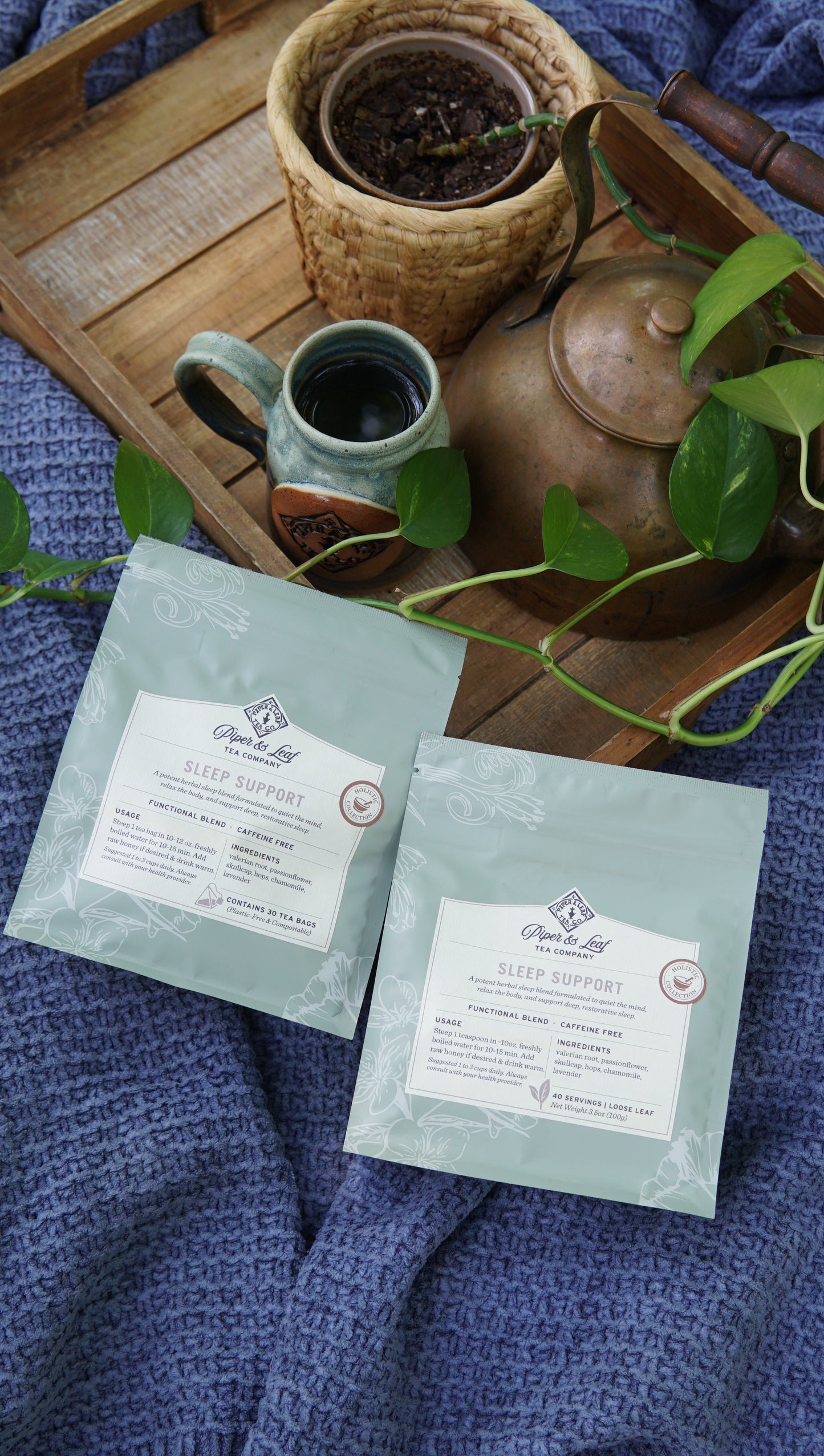 Two pouches of Piper and Leaf Tea Co.'s Sleep Support - Holistic Collection rest on a blue blanket, accompanied by a wooden tray holding a teapot, cup, potted plant, and a green vine.