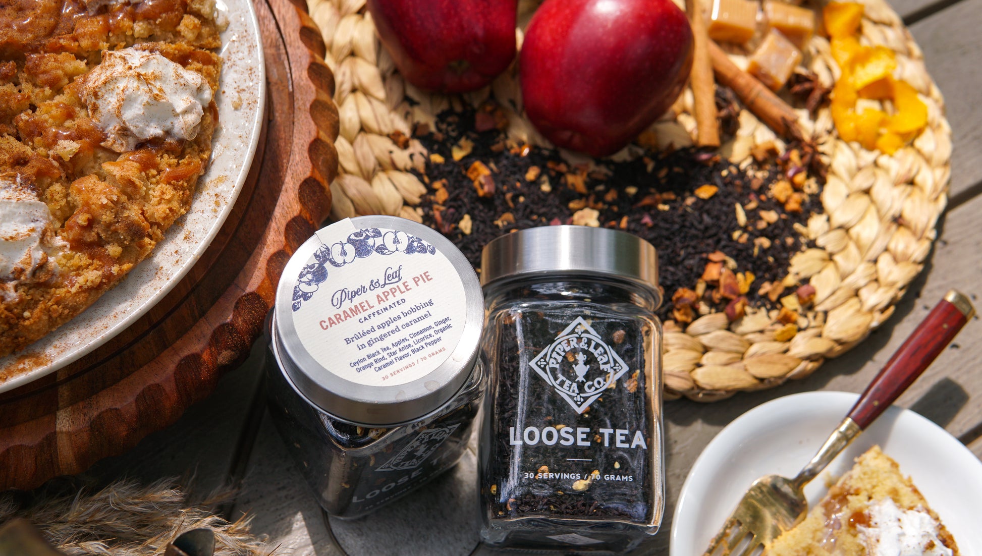 A Caramel Apple Pie Glass Jar of Loose Leaf Tea by Piper & Leaf Tea Co. sits on a woven mat beside apples, caramel, cinnamon sticks, and a caramel apple pie—capturing the essence of this flavorful tea blend.