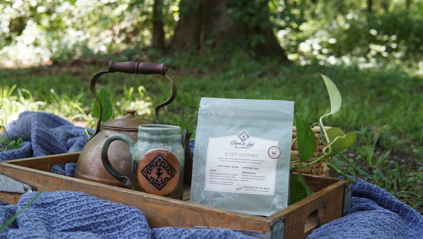 A wooden tray featuring a kettle, a mug, and a Piper and Leaf Tea Co. "Sleep Support - Holistic Collection" bag rests on a blue blanket outdoors, surrounded by greenery in the background.