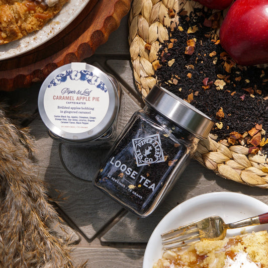 A Piper & Leaf Tea Co. Caramel Apple Pie Glass Jar of Loose Leaf Tea (30 servings) sits on a table with apples, loose tea leaves, and a plate of pie.