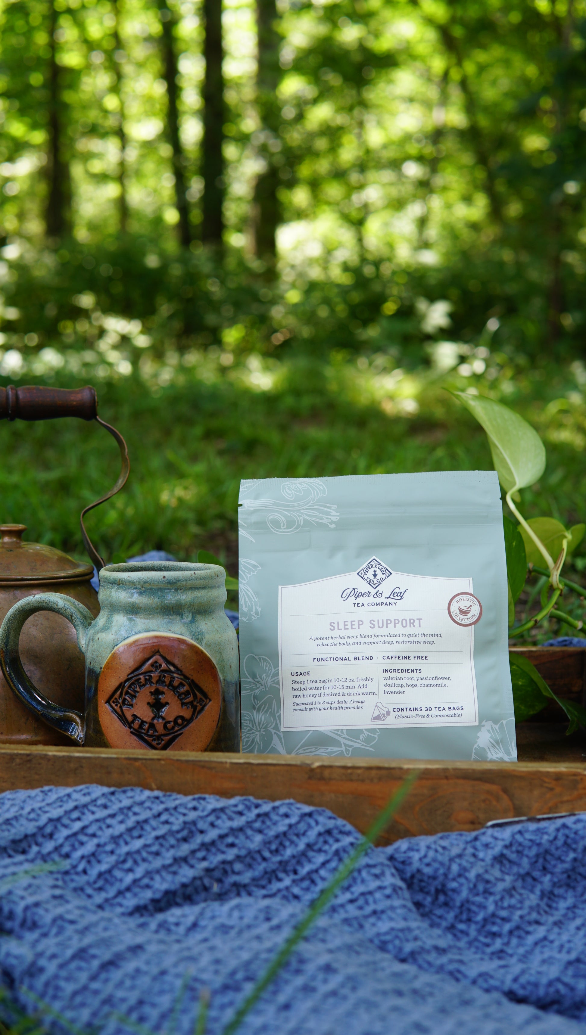A package of Piper and Leaf Tea Co.’s Sleep Support – Holistic Collection herbal tea sits next to a ceramic mug and a blue knitted cloth outdoors, with greenery in the background.