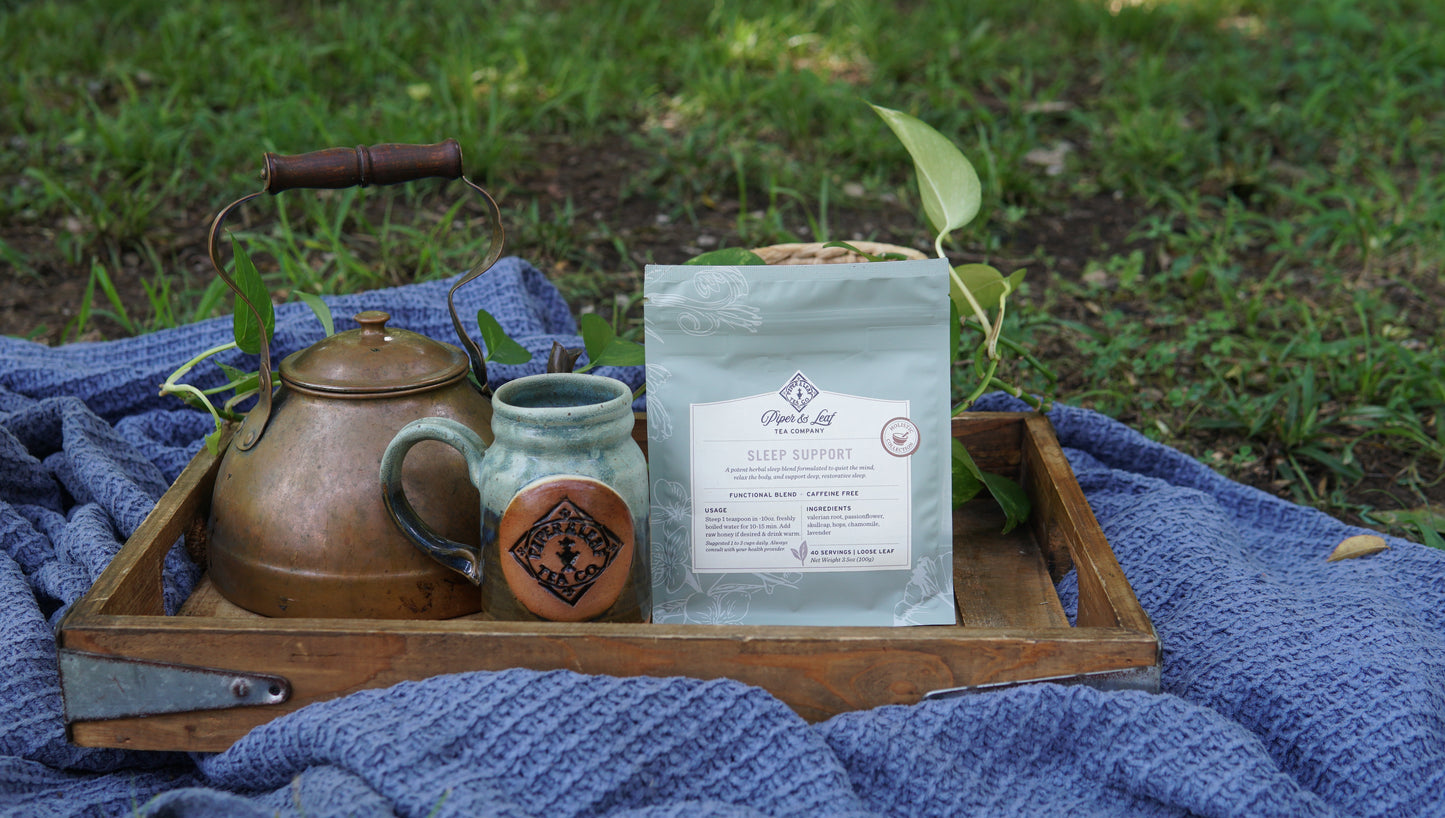 A wooden tray on a blue blanket holds a copper teapot, a ceramic mug, and a Piper and Leaf Tea Co. Sleep Support - Holistic Collection package, set outdoors on the grass.