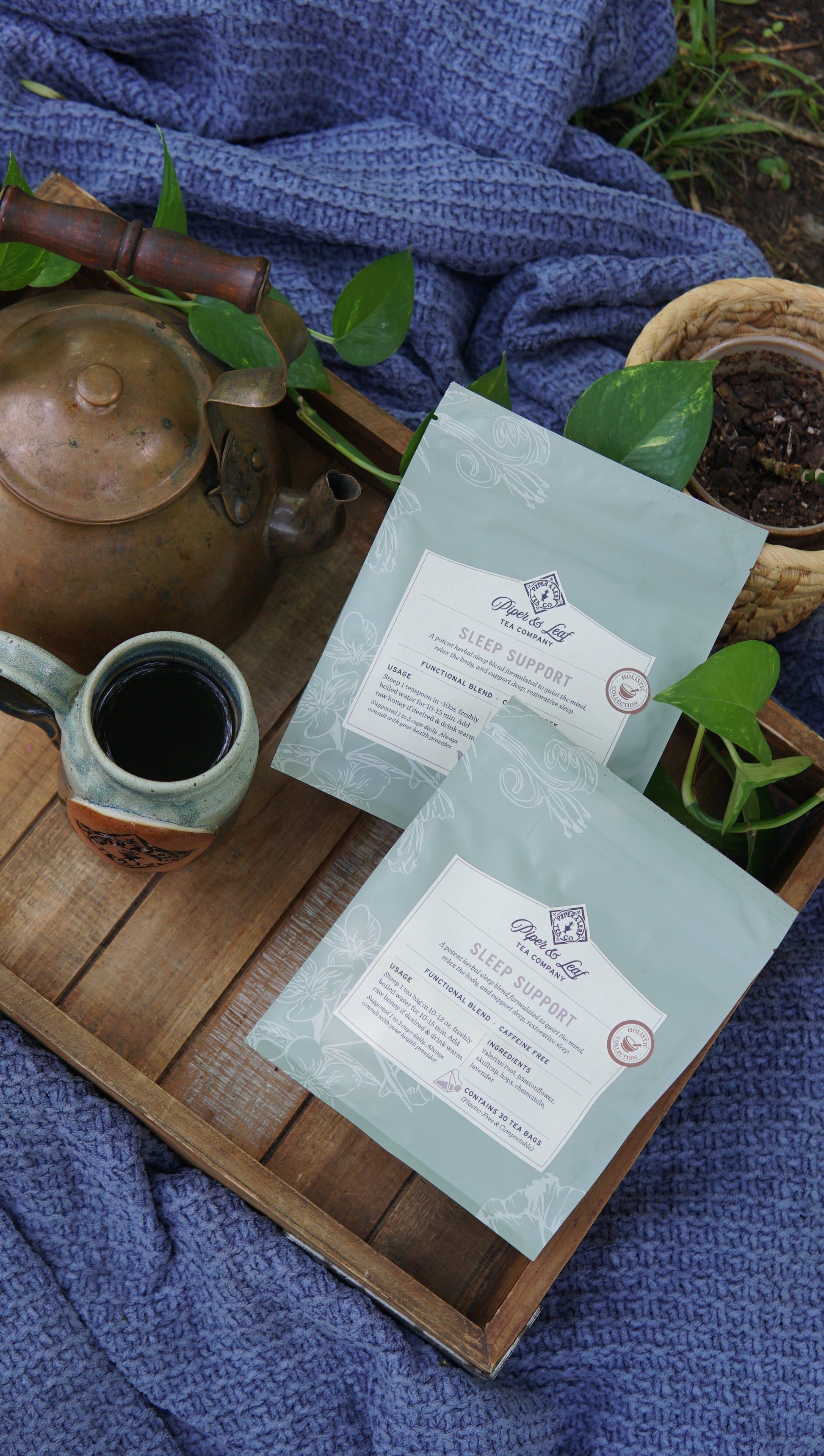 Two light blue packets of Piper and Leaf Tea Co.’s Sleep Support - Holistic Collection are displayed on a wooden tray alongside a ceramic mug, teapot, and green plants, all arranged on a textured blue cloth.