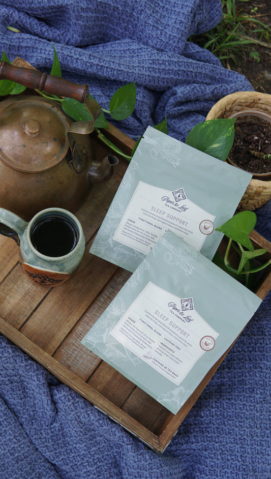 Two light blue packets of Piper and Leaf Tea Co.’s Sleep Support - Holistic Collection are displayed on a wooden tray alongside a ceramic mug, teapot, and green plants, all arranged on a textured blue cloth.