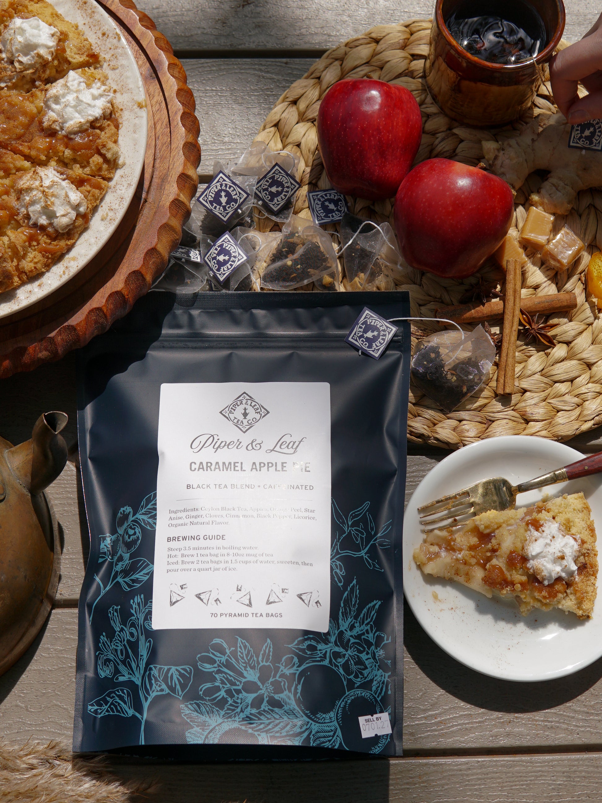 A bag of Piper & Leaf Tea Co. Caramel Apple Pie - 70ct Tea Bags sits on a table with crisp apples, Ceylon black tea bags, a slice of pie, a steaming cup of tea, and cinnamon sticks arranged nearby.