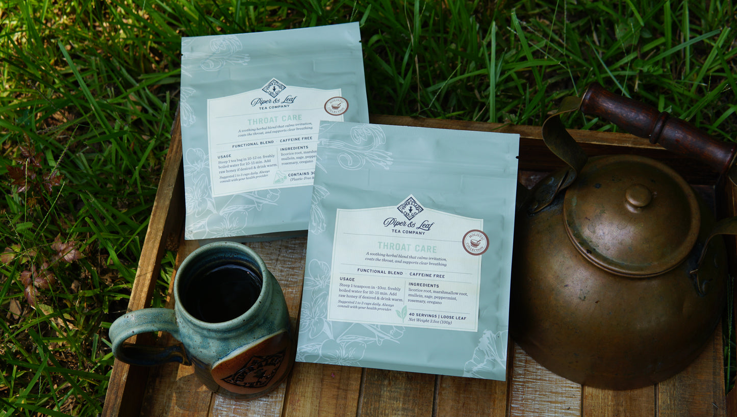 Two packets of Piper and Leaf Tea Co.'s "Throat Care - Holistic Collection" rest on a wooden tray alongside a ceramic mug of tea and a metal teapot, set outdoors on the grass.