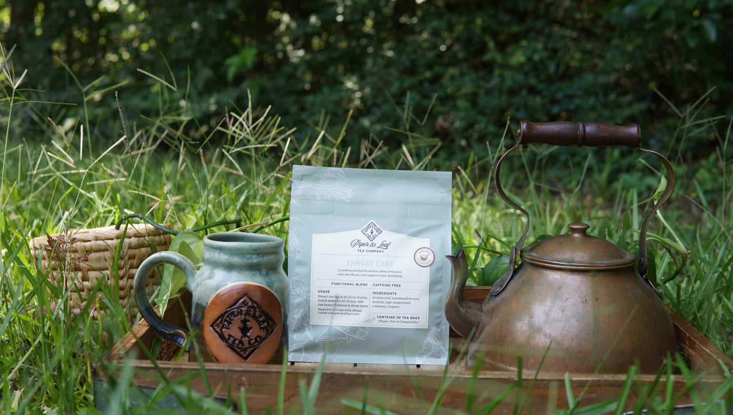 A bag of Piper and Leaf Tea Co.'s "Throat Care - Holistic Collection" tea, accompanied by a ceramic mug, a brass teapot, and a woven basket, are arranged on a wooden tray set in the grass with lush greenery in the background.