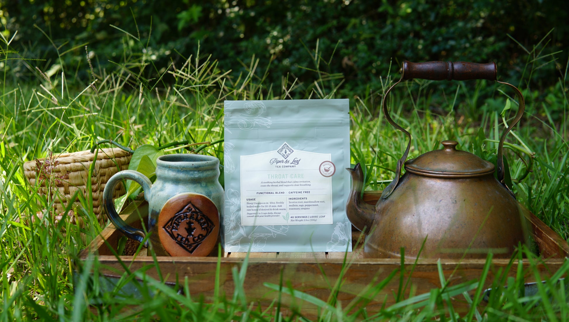 A "Throat Care - Holistic Collection" tea set by Piper and Leaf Tea Co. is arranged on a wooden tray with a mug, tea bag, ear of corn, and metal teapot, all resting on grass amid lush greenery in the background.