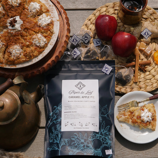 A caramel apple pie with whipped cream, a box of Piper & Leaf Tea Co. Caramel Apple Pie - 70ct Tea Bags, glass of tea, teapot, and wrapped tea bags are displayed on a wooden table.