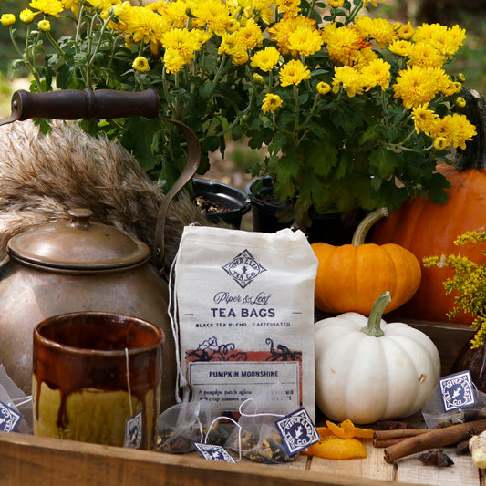 A rustic autumn scene with yellow chrysanthemums, assorted pumpkins, a metal teapot, jar candle, cinnamon sticks, and Piper & Leaf Tea Co. Pumpkin Moonshine 9ct Tea Bags in Muslin on a wooden tray.