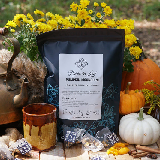 A bag of Piper & Leaf Tea Co. Pumpkin Moonshine Bulk Sachets (70ct tea bags) is displayed outdoors alongside tea bags, a ceramic mug, pumpkins, yellow flowers, and an antique teapot.
