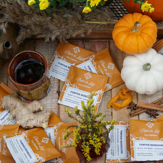 Pumpkin and white gourds, ginger, a cup of tea, and Piper & Leaf Tea Co.'s Pumpkin Moonshine Case of Individually Wrapped Tea Bags (50ct) are displayed on a rustic wooden surface with flowers and autumn decor.