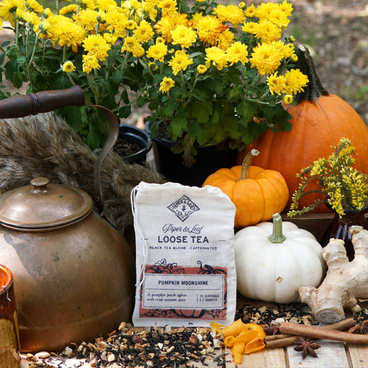 A Piper & Leaf Tea Co. Pumpkin Moonshine Muslin Bag of Loose Leaf Tea (15 servings) rests on a wooden table with yellow flowers, pumpkins, a brass kettle, and scattered tea leaves in an outdoor autumn spice setting.