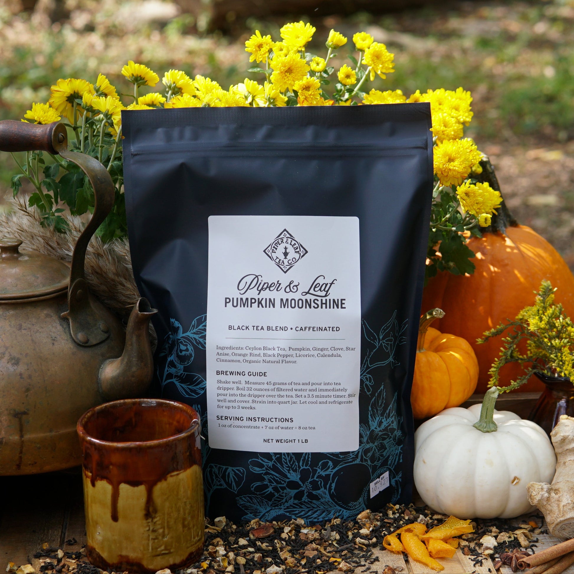 A black Piper & Leaf Tea Co. Pumpkin Moonshine Pound Bag (190 servings) is shown outdoors among yellow flowers, pumpkins, a mug, a brass teapot, and autumn decorations.