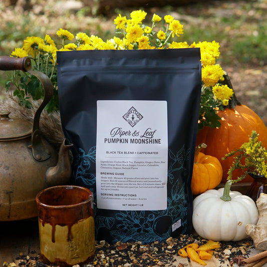 A black Piper & Leaf Tea Co. Pumpkin Moonshine Pound Bag (190 servings) is shown outdoors among yellow flowers, pumpkins, a mug, a brass teapot, and autumn decorations.