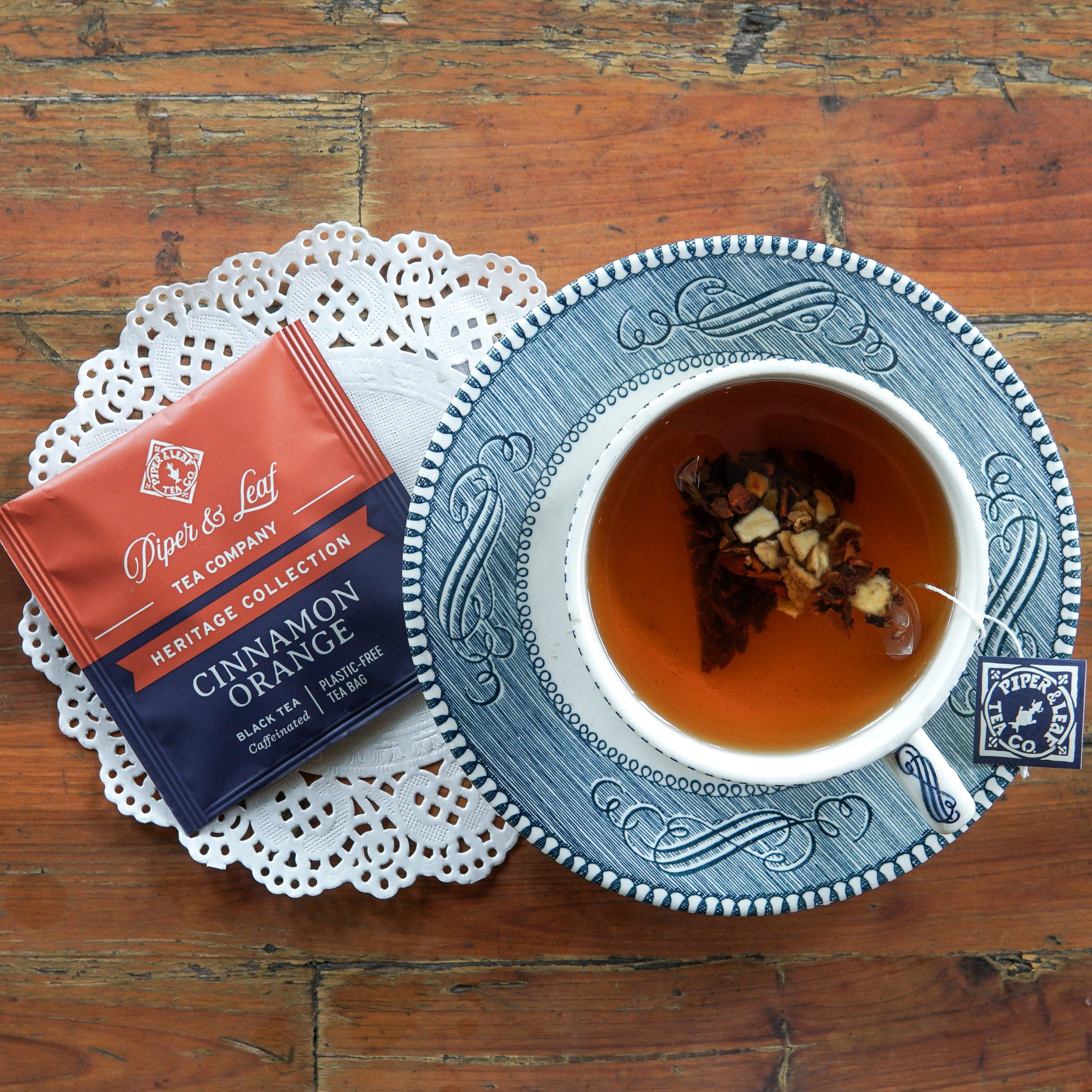 A cup of tea with floating spices rests on a blue saucer beside a box of Piper and Leaf Tea Co. Cinnamon Orange 50ct Individually Wrapped Tea Bags, set on a doily atop a wooden table.