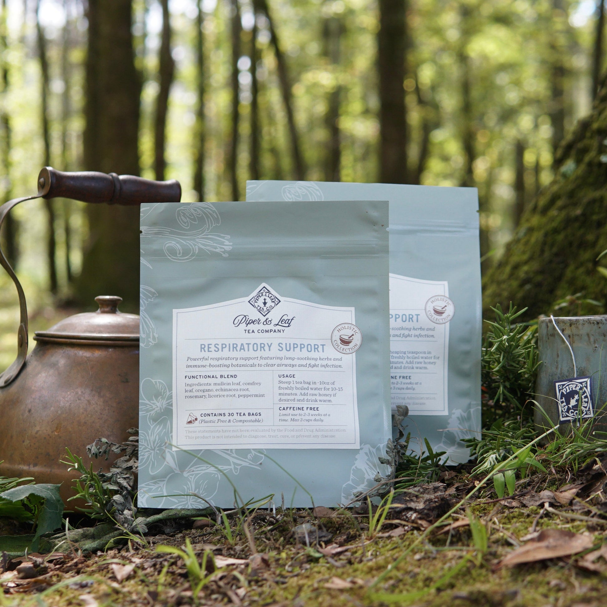 Two bags of Piper and Leaf Tea Co.’s Respiratory Support tea sit on moss in a forest next to a metal teapot and tea bag, surrounded by trees—an inviting display of soothing herbal support for easy breathing.