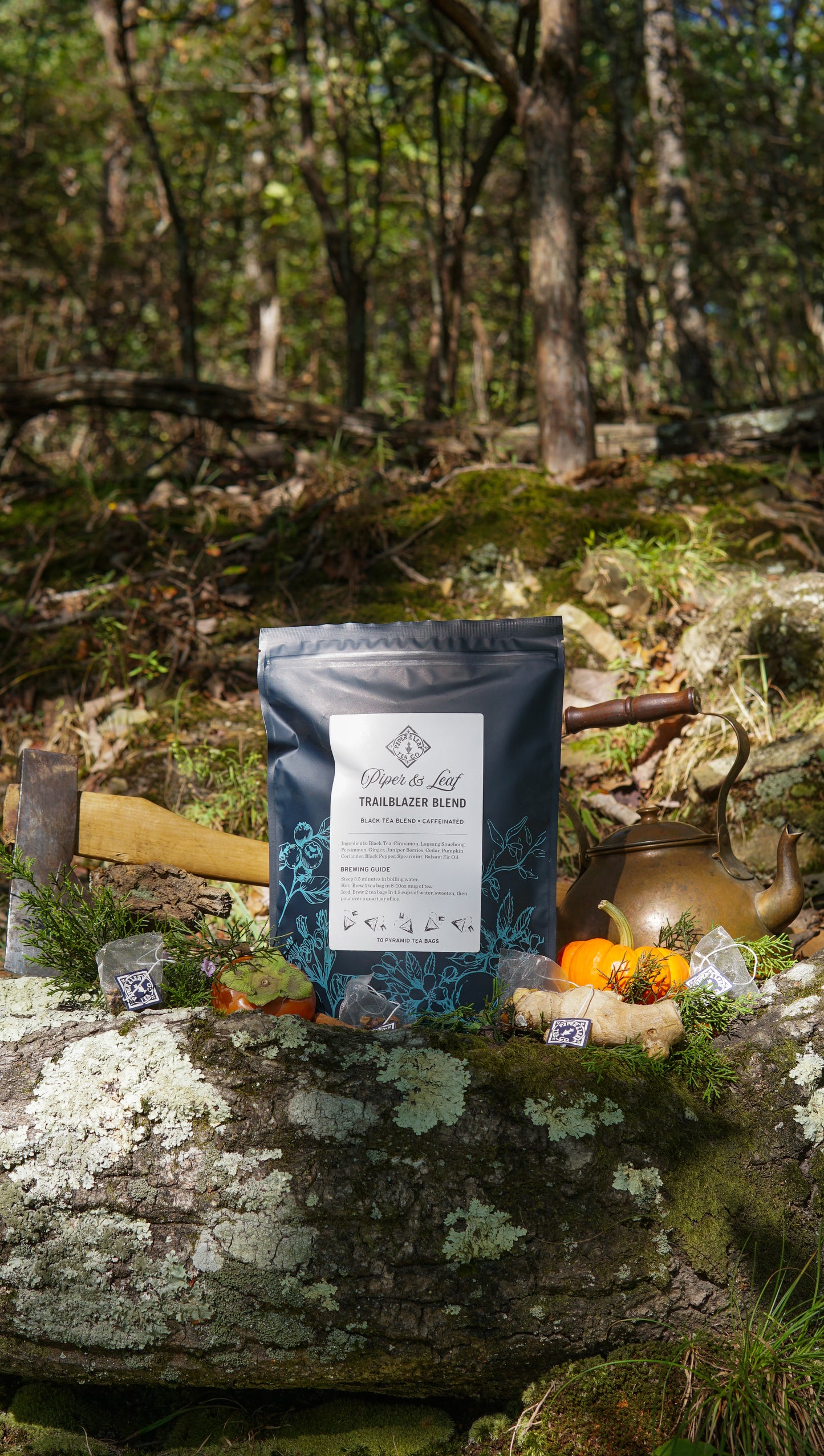 A box of Piper & Leaf Tea Co.'s Trailblazer Blend Bulk Sachets (70ct Tea Bags) rests on a mossy log outdoors, surrounded by an axe, teapot, pumpkins, and packages against a peaceful forest backdrop.