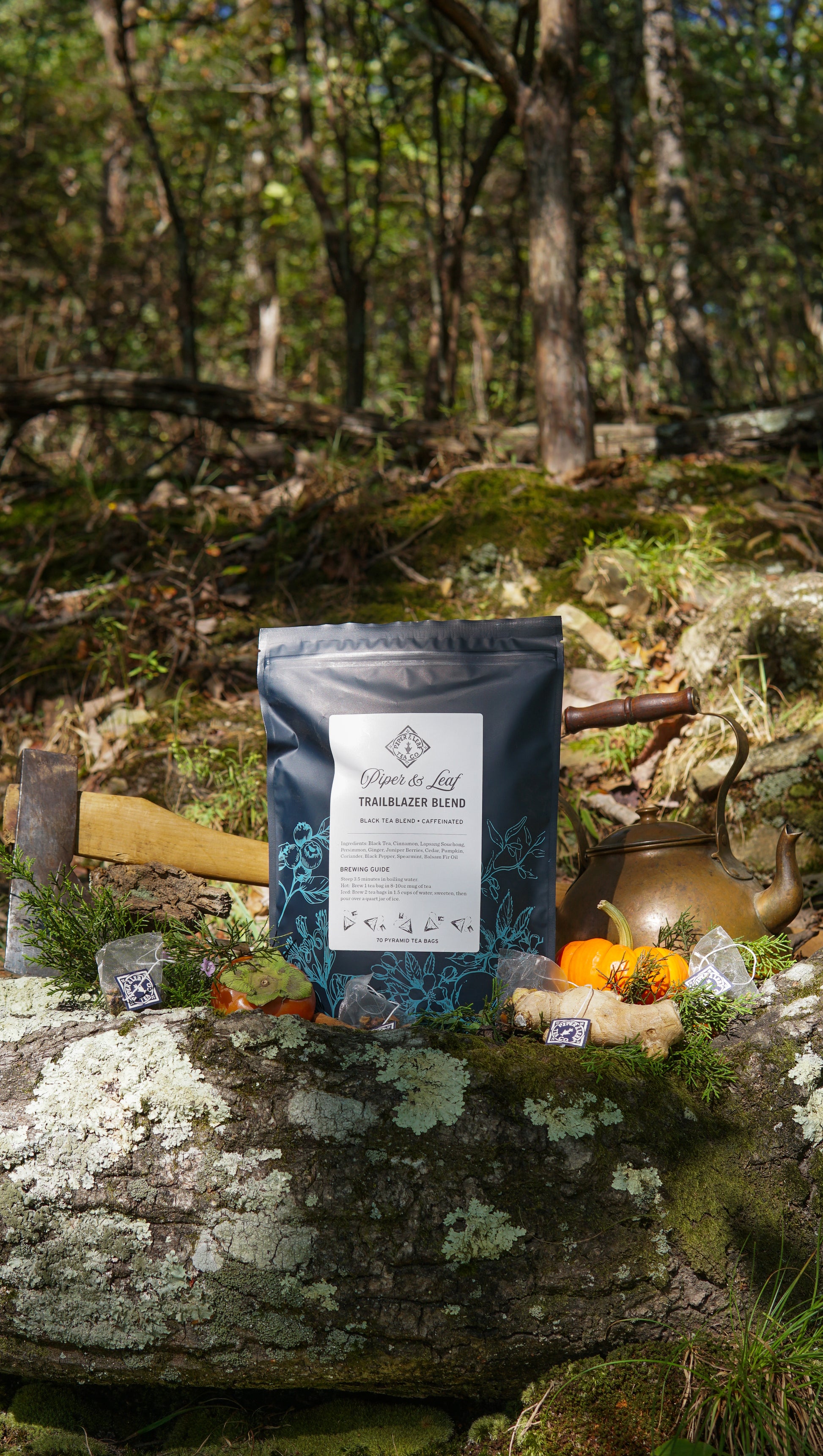A box of Piper & Leaf Tea Co.'s Trailblazer Blend Bulk Sachets (70ct Tea Bags) rests on a mossy log outdoors, surrounded by an axe, teapot, pumpkins, and packages against a peaceful forest backdrop.