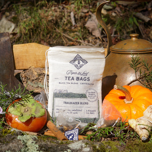 A bag of Piper & Leaf Tea Co. Trailblazer Blend 9ct Tea Bags in Muslin is displayed outdoors with a persimmon, pumpkin, herbs, a brass teapot, cinnamon sticks, and extra tea bags.