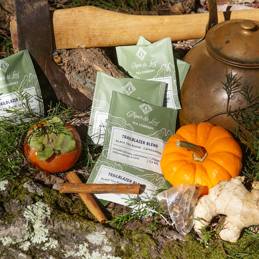 Piper & Leaf Tea Co.'s Trailblazer Blend Case of Individually Wrapped Tea Bags (50ct) is displayed outdoors with an axe, kettle, mini pumpkin, persimmon, cinnamon sticks, and greenery on a mossy log.