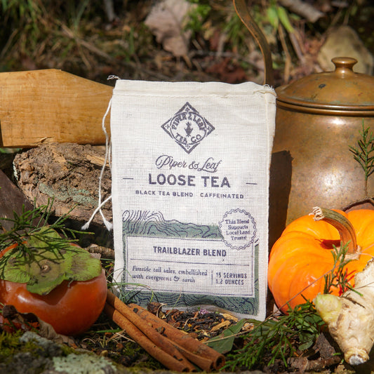 A Piper & Leaf Tea Co. Trailblazer Blend Muslin Bag of Loose Leaf Tea (15 servings) rests outdoors with a persimmon, pumpkin, cinnamon sticks, and brass teapot—creating an ideal autumn setting for this smoky tea blend.