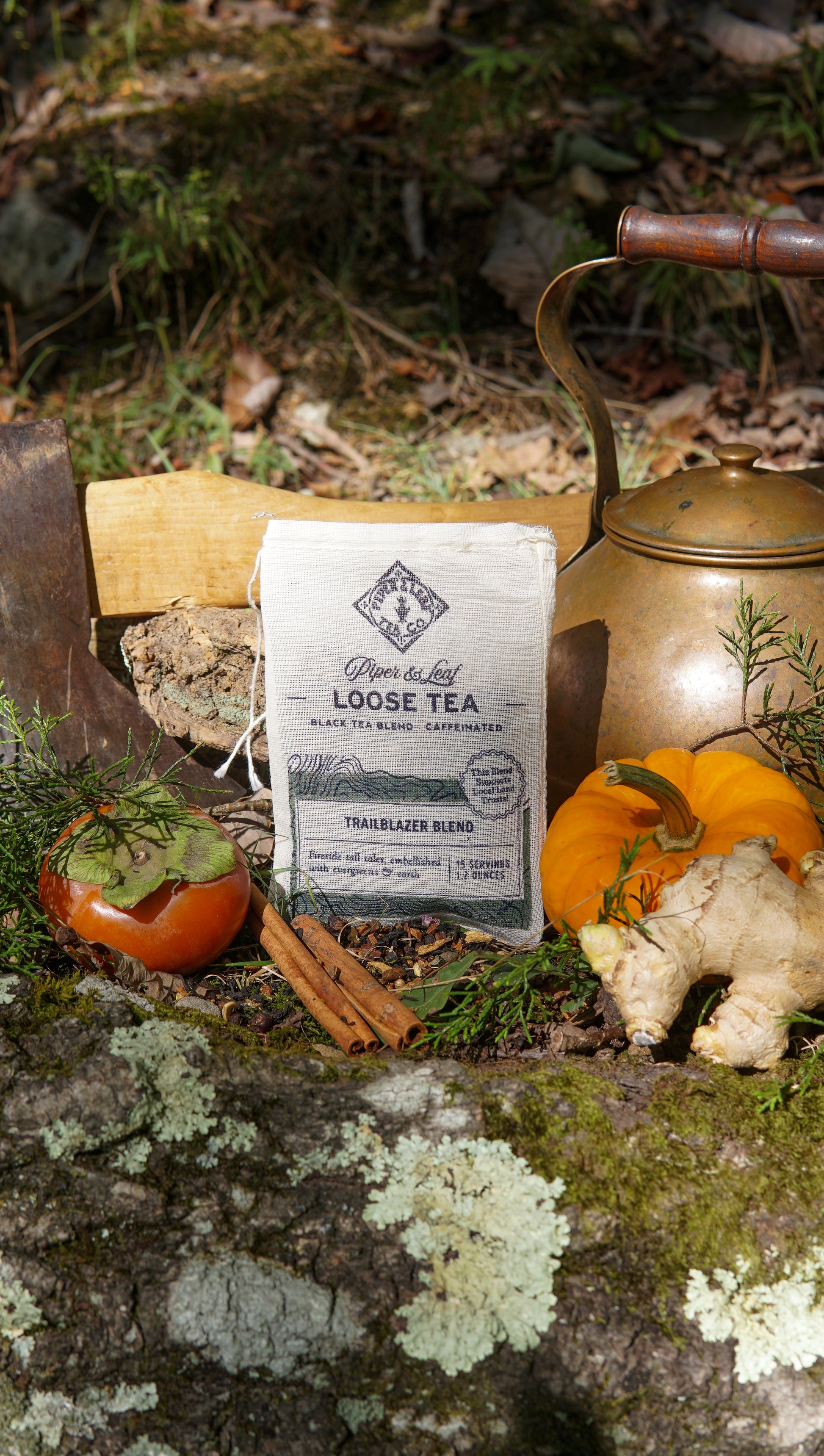 A Trailblazer Blend Muslin Bag of Loose Leaf Tea by Piper & Leaf Tea Co. rests on a mossy rock beside a brass kettle, persimmon, ginger, cinnamon sticks, pumpkin, and wood with greenery and the forest floor in the background.