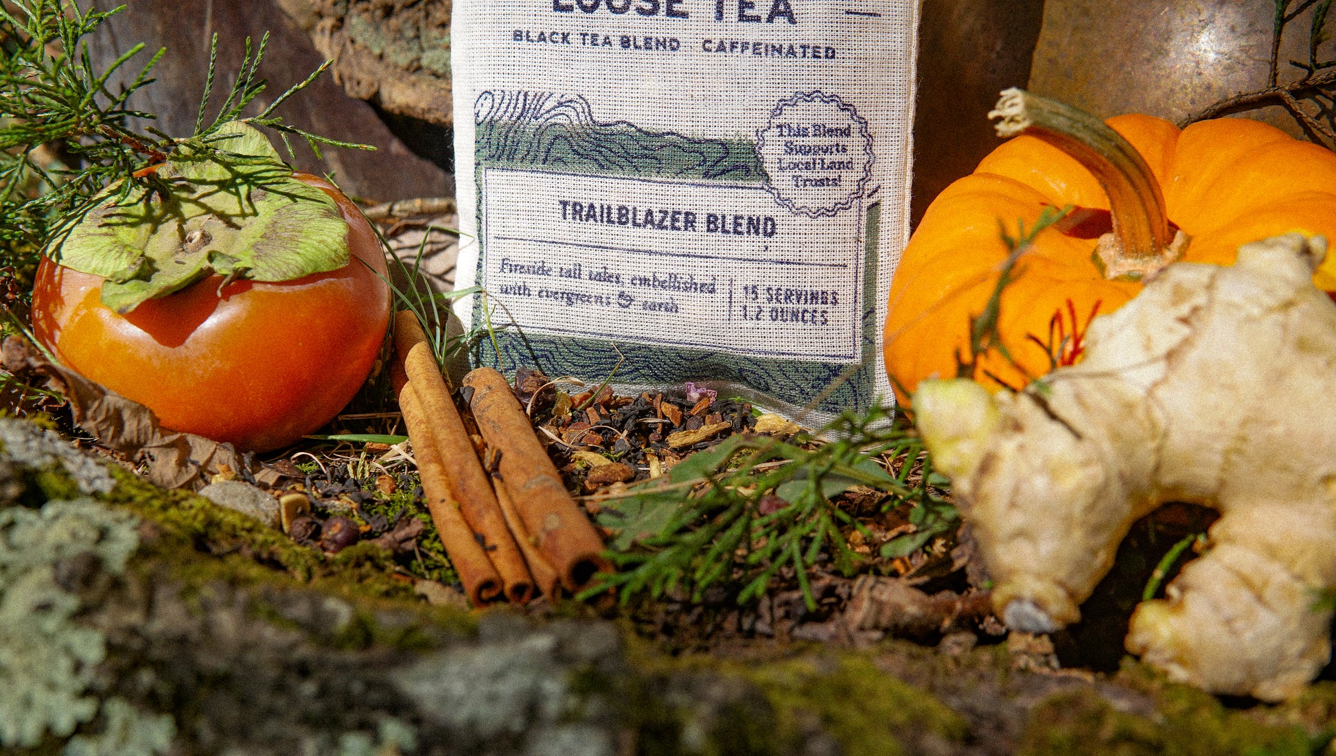 A Trailblazer Blend Muslin Bag of Loose Leaf Tea by Piper & Leaf Tea Co. sits outdoors among a persimmon, two cinnamon sticks, a small pumpkin, ginger root, and green leaves.