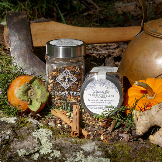 A Piper & Leaf Tea Co. Trailblazer Blend Glass Jar of Loose Leaf Tea (30 servings), cinnamon sticks, a persimmon, small pumpkin, axe, and kettle are displayed on a mossy log in the outdoors.