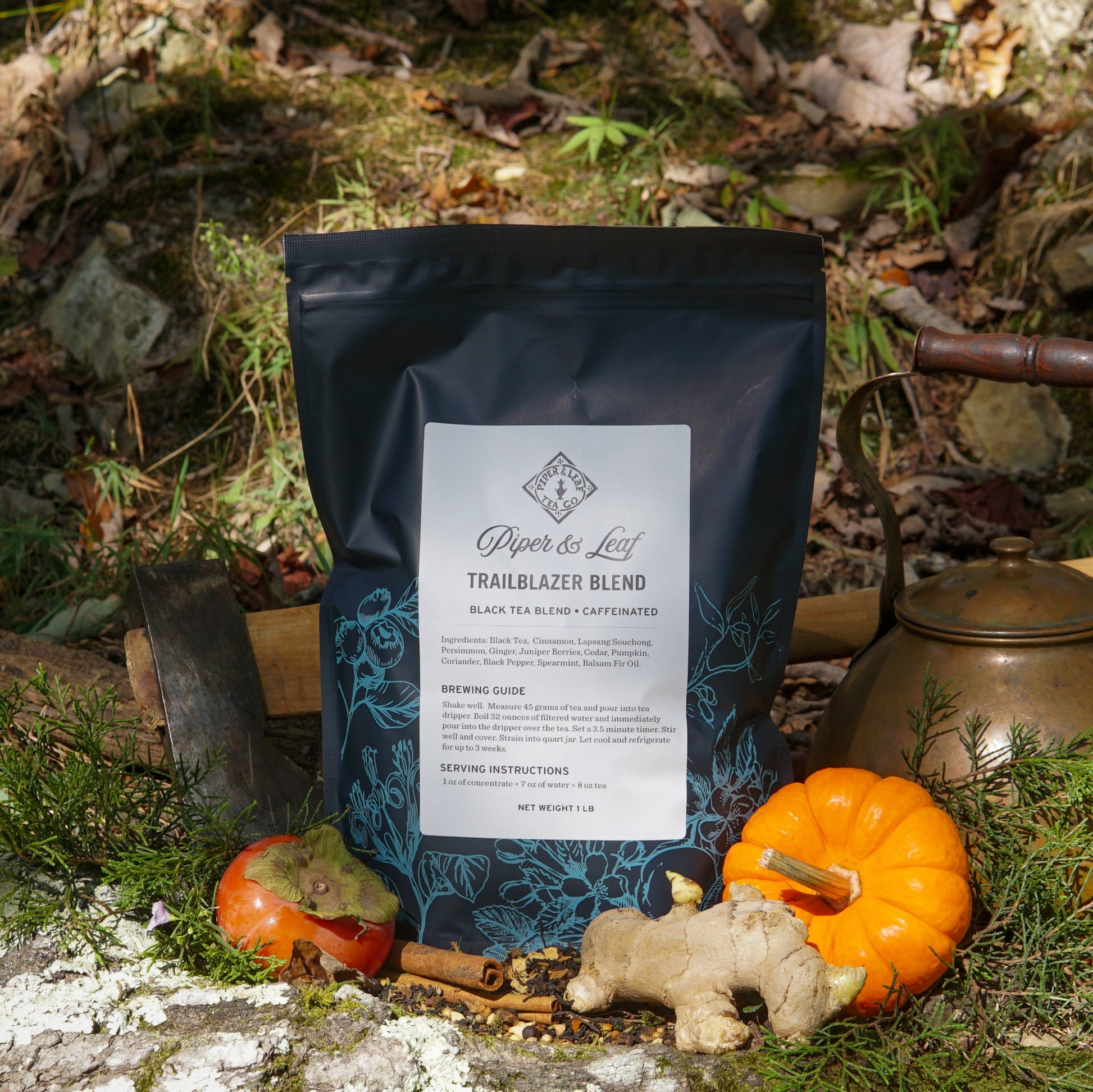 A Piper & Leaf Tea Co. Trailblazer Blend Pound Bag (190 servings) is displayed outdoors on rocks with an axe, brass kettle, orange pumpkin, ginger root, and apple among greenery.