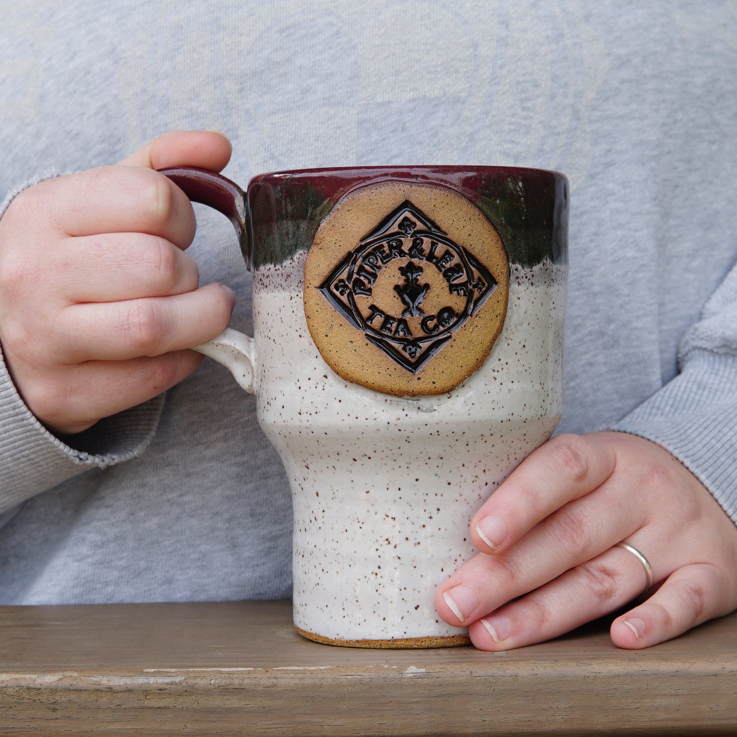 A person wearing a gray sweater holds a large Pottery Travel Mug with a “Piper and Leaf Tea Co.” logo while resting it on a wooden surface.