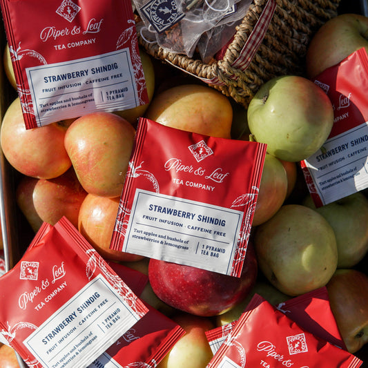 A case of 50 Strawberry Shindig Tea Bag Envelopes by Piper & Leaf Tea Co. sits atop a pile of apples, with a woven basket visible in the corner.