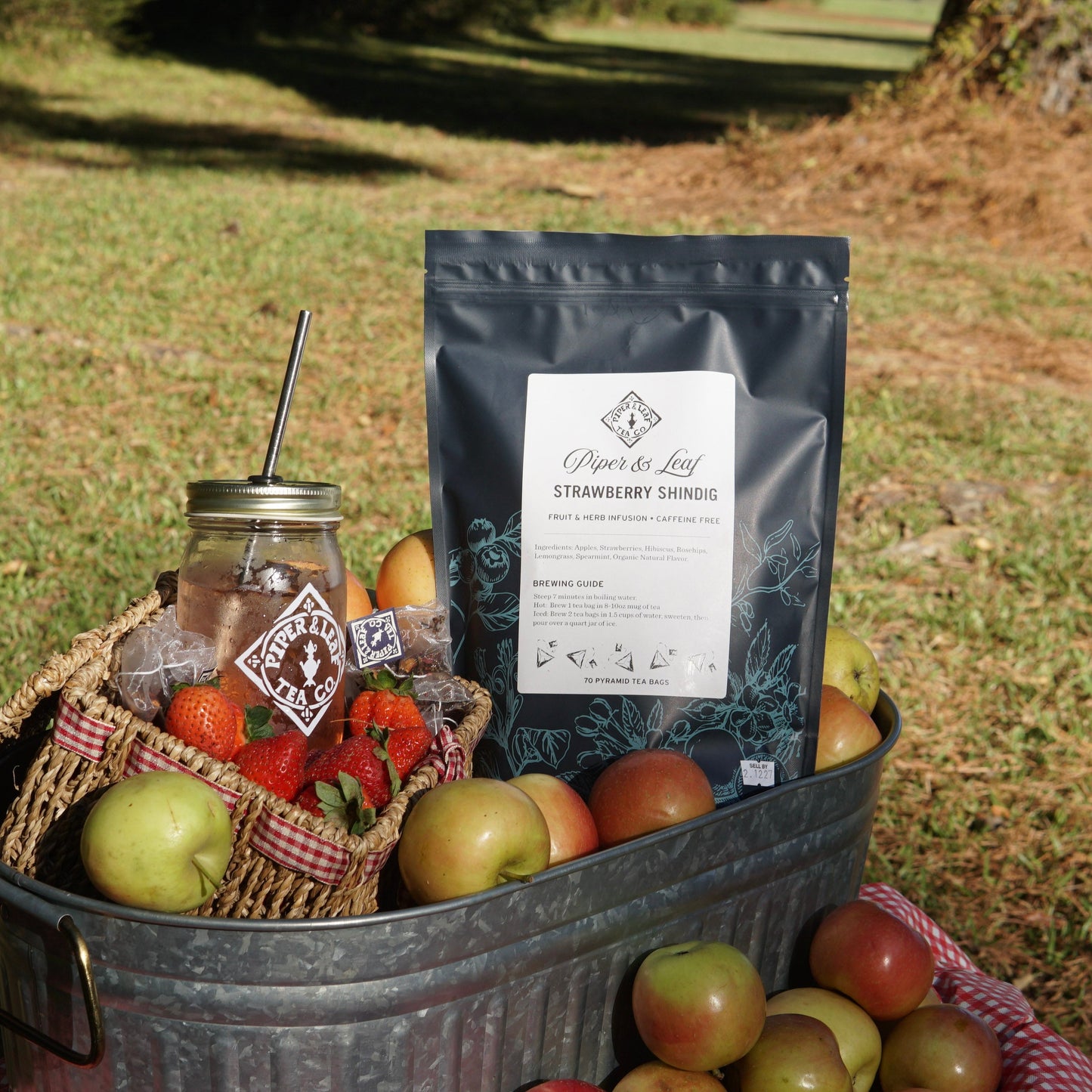 A package of Piper & Leaf Tea Co. Strawberry Shindig Bulk Sachets (70ct Tea Bags), a mason jar of iced tea, apples, and strawberries are displayed in a metal tub outside on a sunny day.