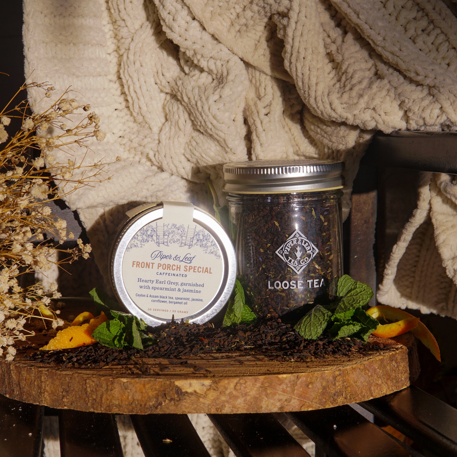 A tin and a Piper & Leaf Tea Co. Front Porch Special Glass Jar of Loose leaf Tea (30 servings) rest on a wooden slab, with dried leaves, orange peel, and a cream knit blanket in the background.