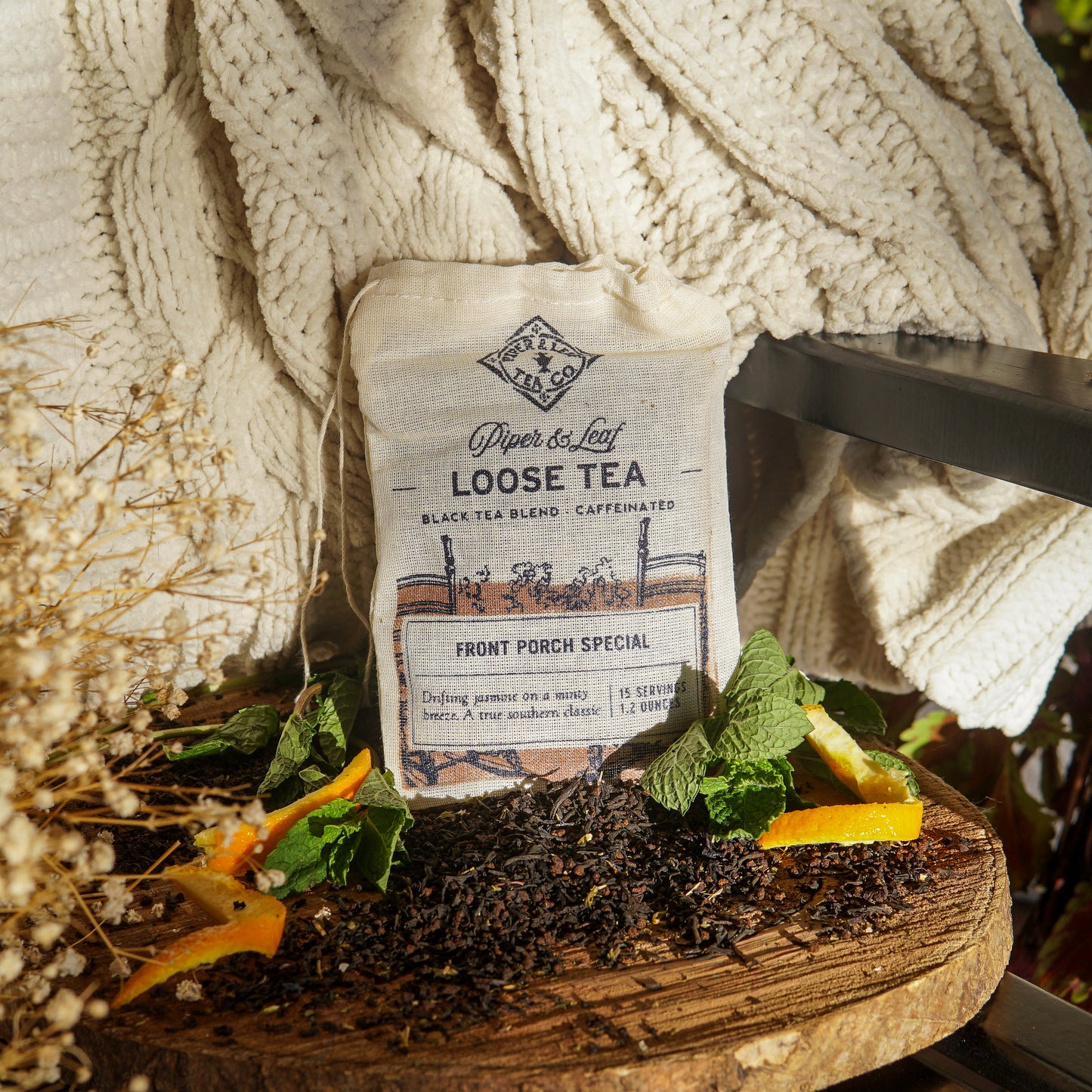 A Piper & Leaf Tea Co. Front Porch Special Muslin Bag of Loose Leaf Tea (15 servings) sits on wood with tea leaves, mint, and orange slices, next to a cream blanket in sunlight—a Southern Living Food Award winner.