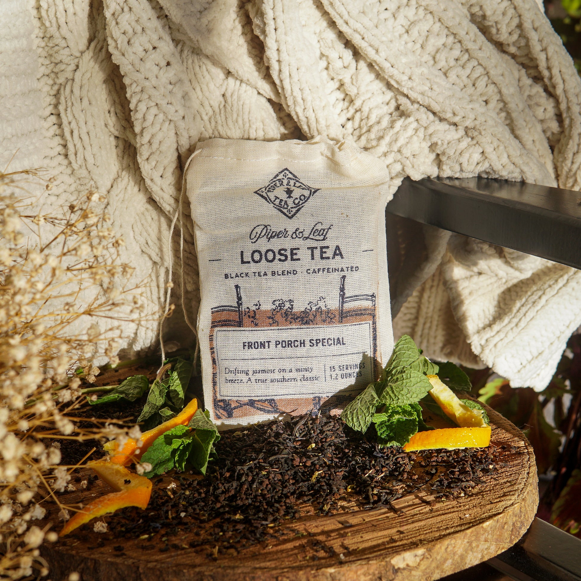 A Piper & Leaf Tea Co. Front Porch Special Muslin Bag of Loose Leaf Tea (15 servings) sits on wood with tea leaves, mint, and orange slices, next to a cream blanket in sunlight—a Southern Living Food Award winner.