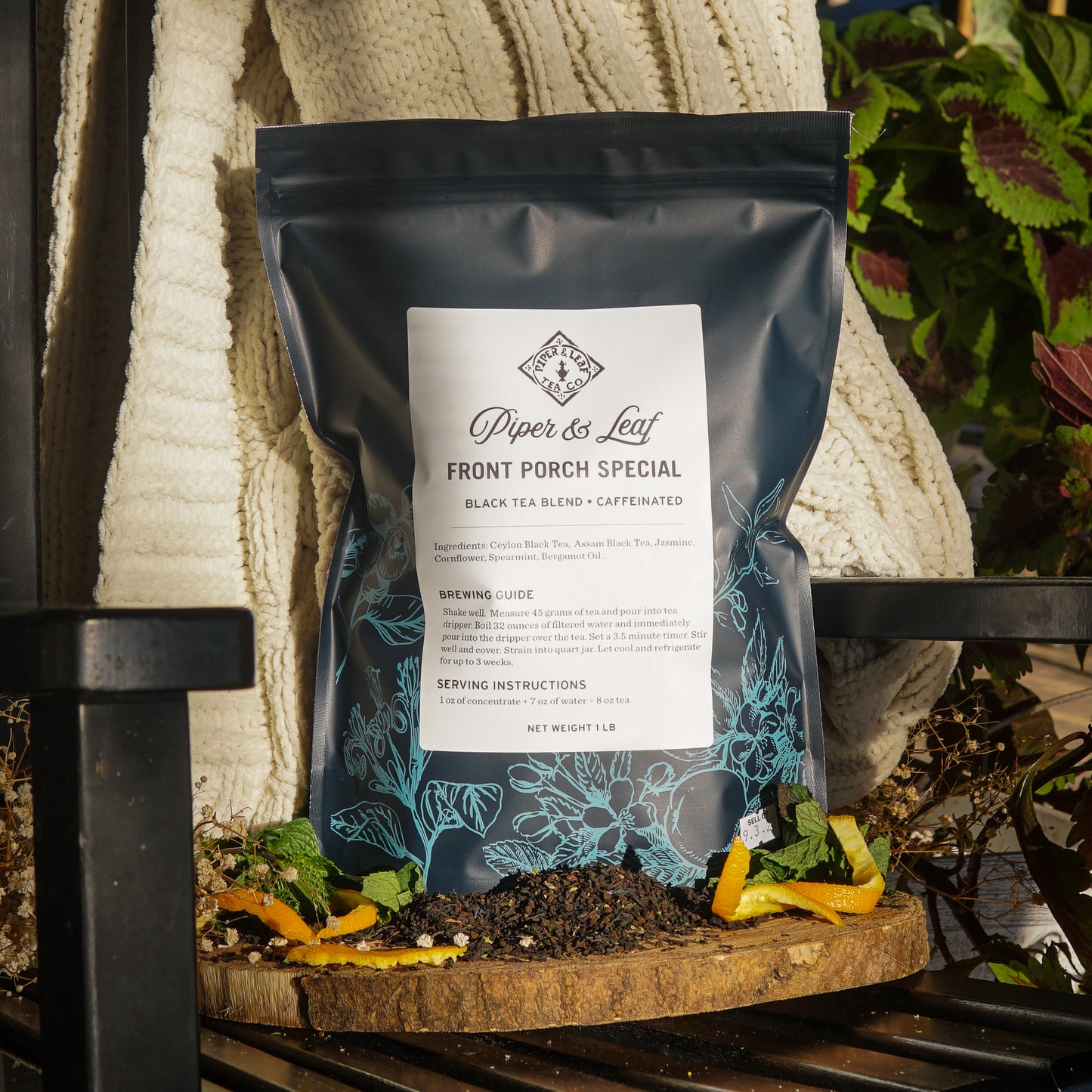 A Piper & Leaf Tea Co. Front Porch Special Pound Bag (190 servings) sits on a wooden board with scattered tea leaves, orange peel, and mint, set against a cozy knitted blanket backdrop.