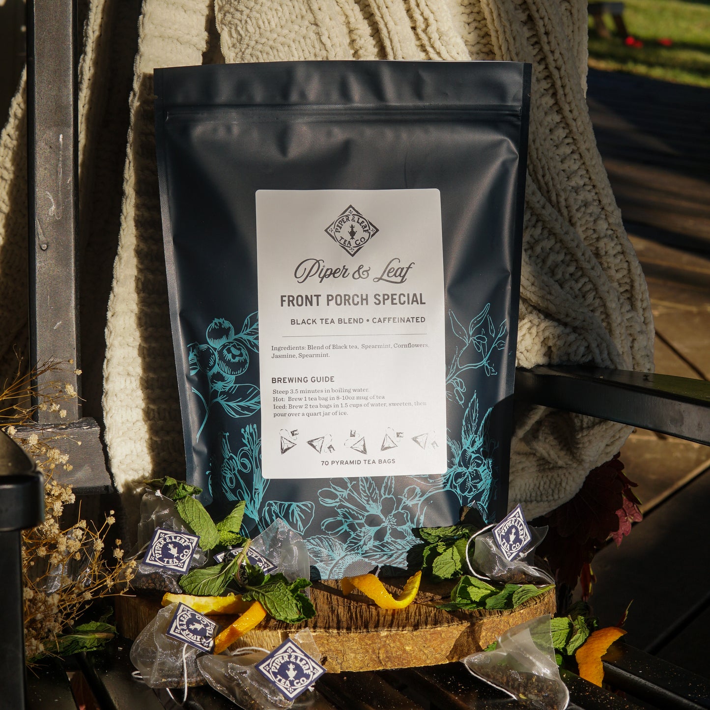 A box of Piper & Leaf Tea Co. Front Porch Special Bulk Sachets - 70ct Tea Bags sits on a bench with mint leaves, orange peels, loose tea, and a knitted blanket, highlighting the comforting, minty jasmine-inspired blend.