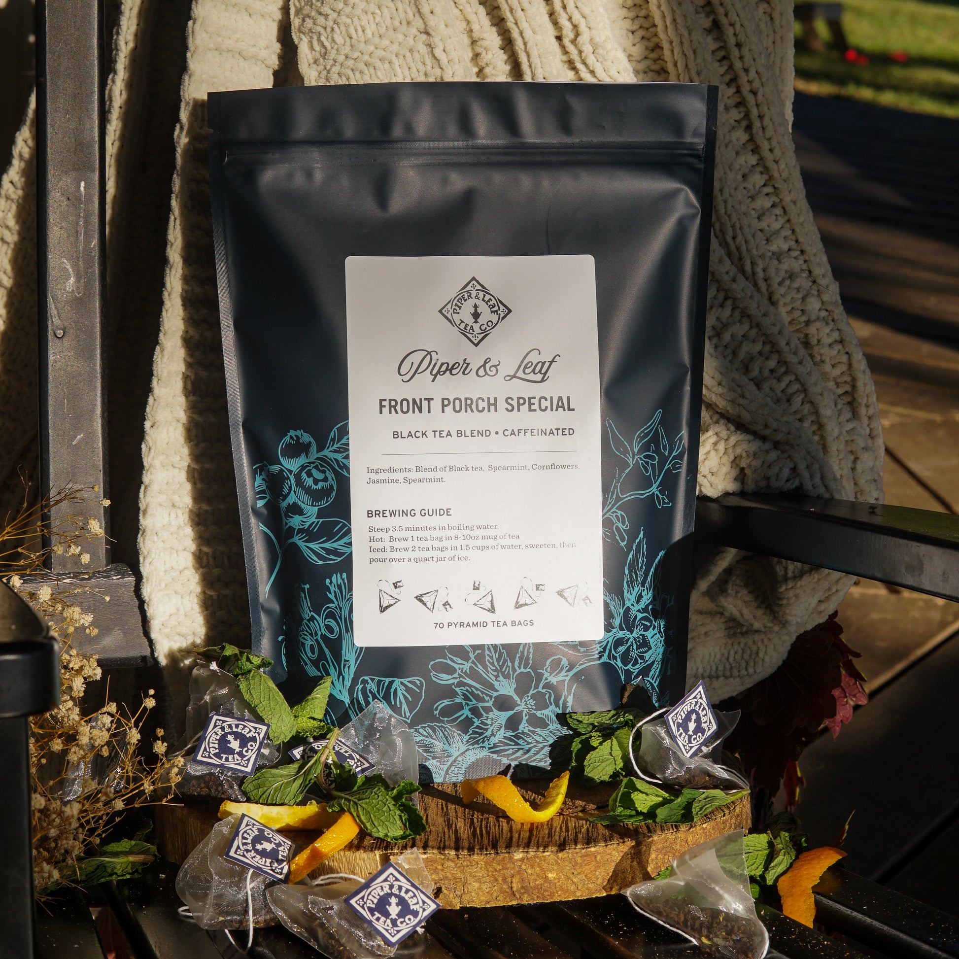 A box of Piper & Leaf Tea Co. Front Porch Special Bulk Sachets - 70ct Tea Bags sits on a bench with mint leaves, orange peels, loose tea, and a knitted blanket, highlighting the comforting, minty jasmine-inspired blend.