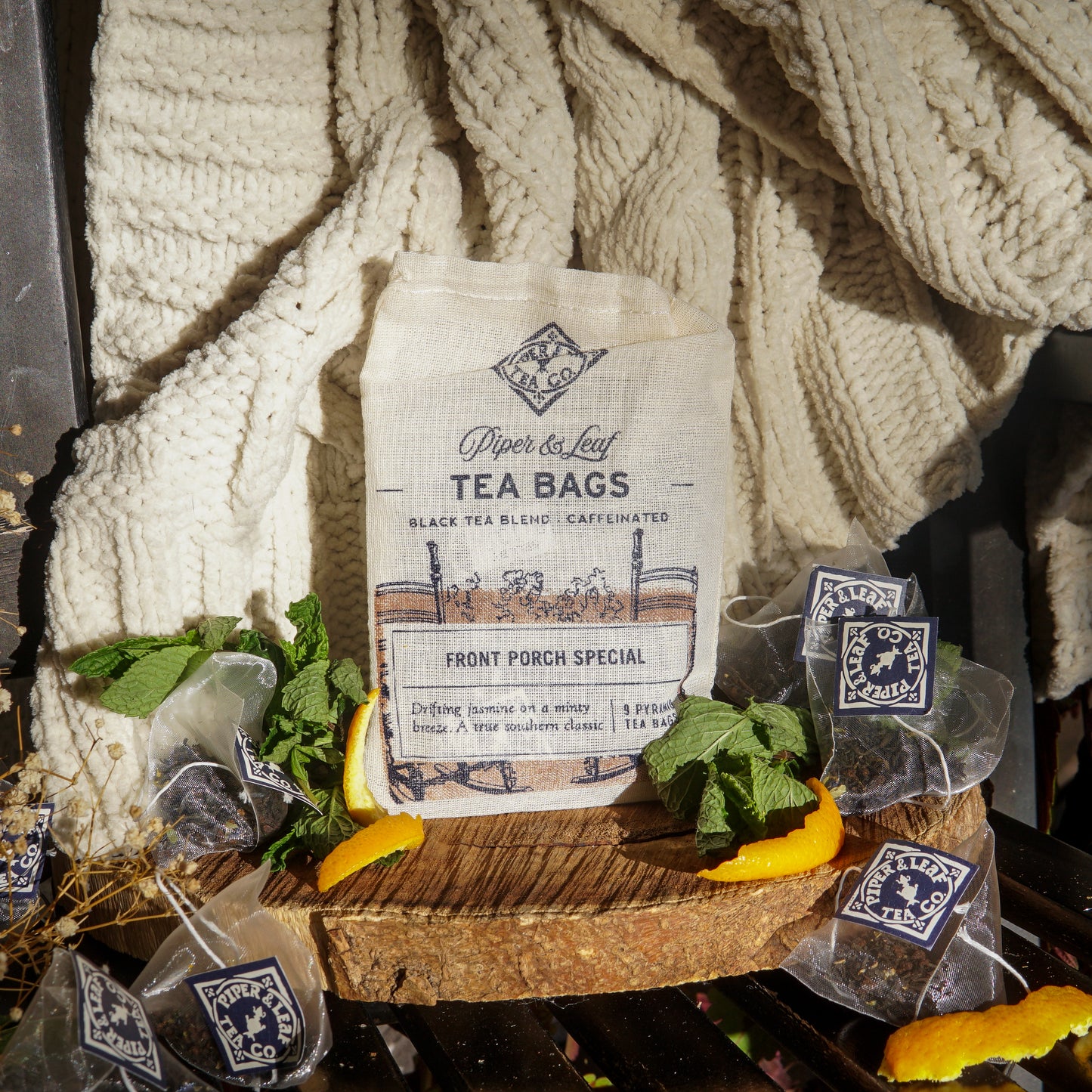 A bag of Piper & Leaf Tea Co. Front Porch Special 9ct Tea Bags in Muslin sits on a knit blanket, surrounded by pyramid sachets, hand-printed muslin bags, fresh mint, and orange slices.