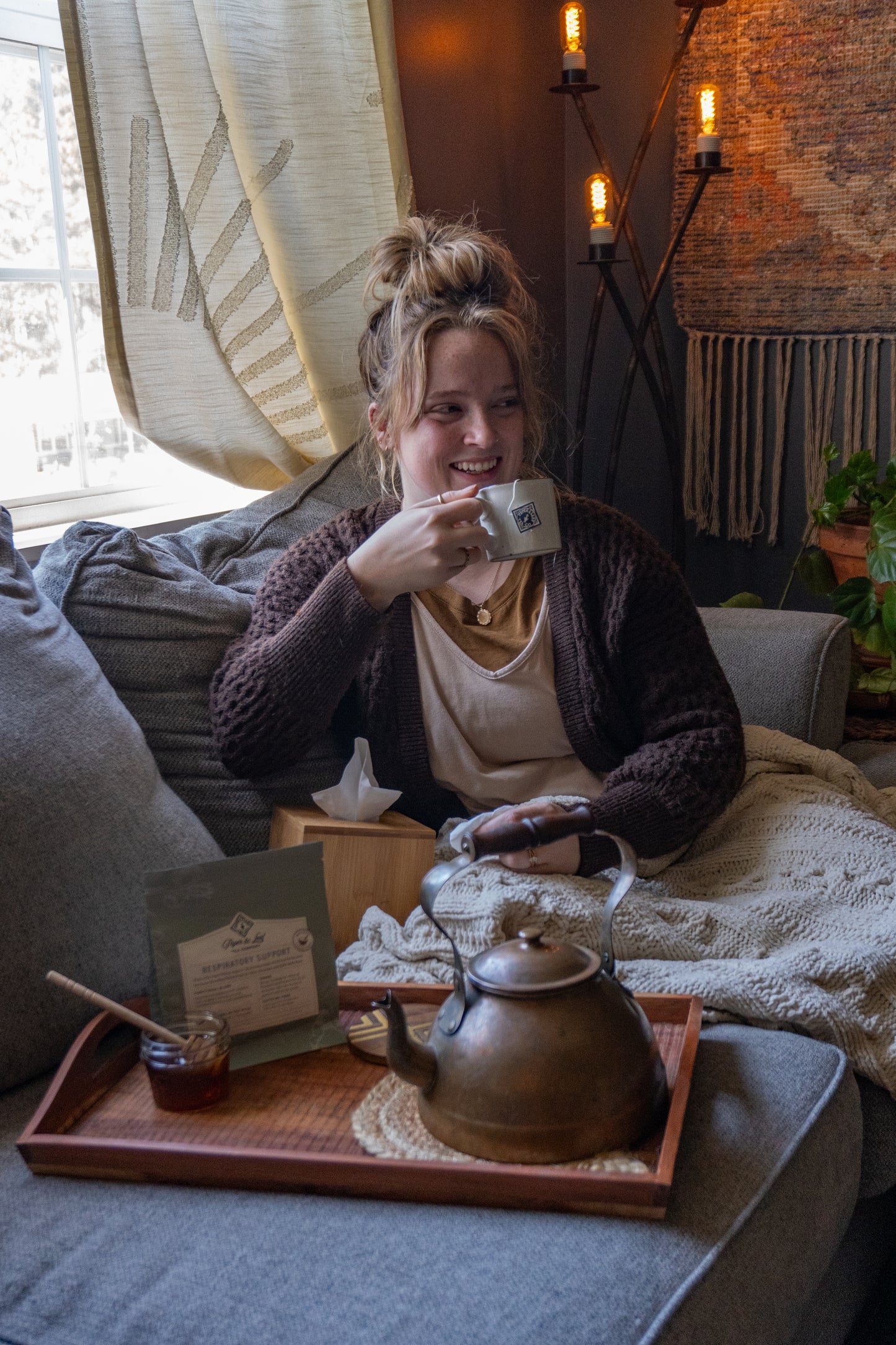A woman sits on a couch by a window, smiling with a cup in hand. Beside her is a tray with Piper and Leaf Tea Co.’s Respiratory Support herbal tea, honey, and a teapot on a blanket. Warm lighting highlights the cozy decor.