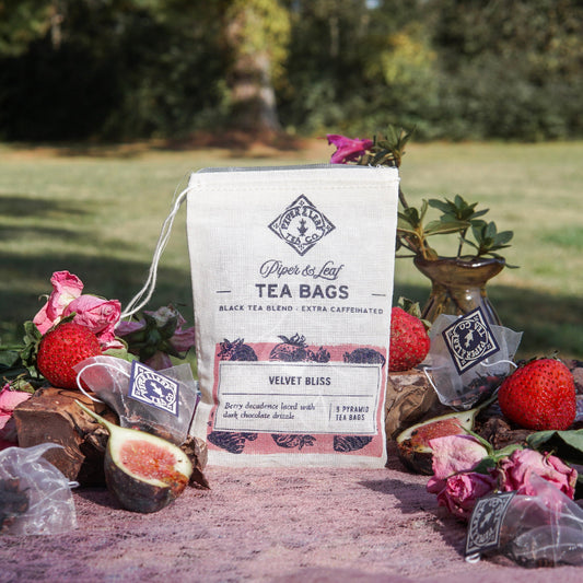 A bag of Piper & Leaf Tea Co. Velvet Bliss 9ct Tea Bags in Muslin rests on a pink surface outdoors, surrounded by strawberries, figs, rose petals, and creamy chocolate tea bags, with lush greenery in the background.